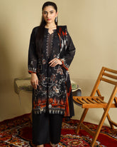 A woman models a three-piece black ethnic suit set featuring a printed kurta, wide-leg trousers, and a matching dupatta, styled in a room with a wooden chair and patterned rug.