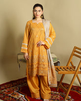 A woman models a mustard yellow ethnic three-piece suit with matching pants and a beige dupatta, standing on a red patterned rug next to a wooden chair against a beige background.