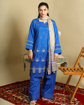 A woman modeling a royal blue three-piece traditional outfit with intricate silver embroidery, matching blue pants, and a light beige patterned scarf, standing on a red patterned rug next to a wooden chair.