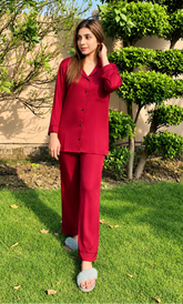 A woman wearing a matching red pajama set with long-sleeve button-up top and wide-leg pants, standing on a green lawn outdoors with trees and a beige wall in the background.