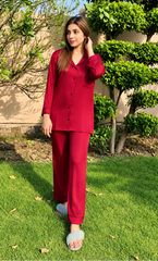 A woman wearing a matching red pajama set with long-sleeve button-up top and wide-leg pants, standing on a green lawn outdoors with trees and a beige wall in the background.