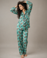 A woman modeling a two-piece pajama set with a long-sleeve button-up top and matching pants, featuring a mint green base with a red and orange floral pattern, set against a plain gray studio background.