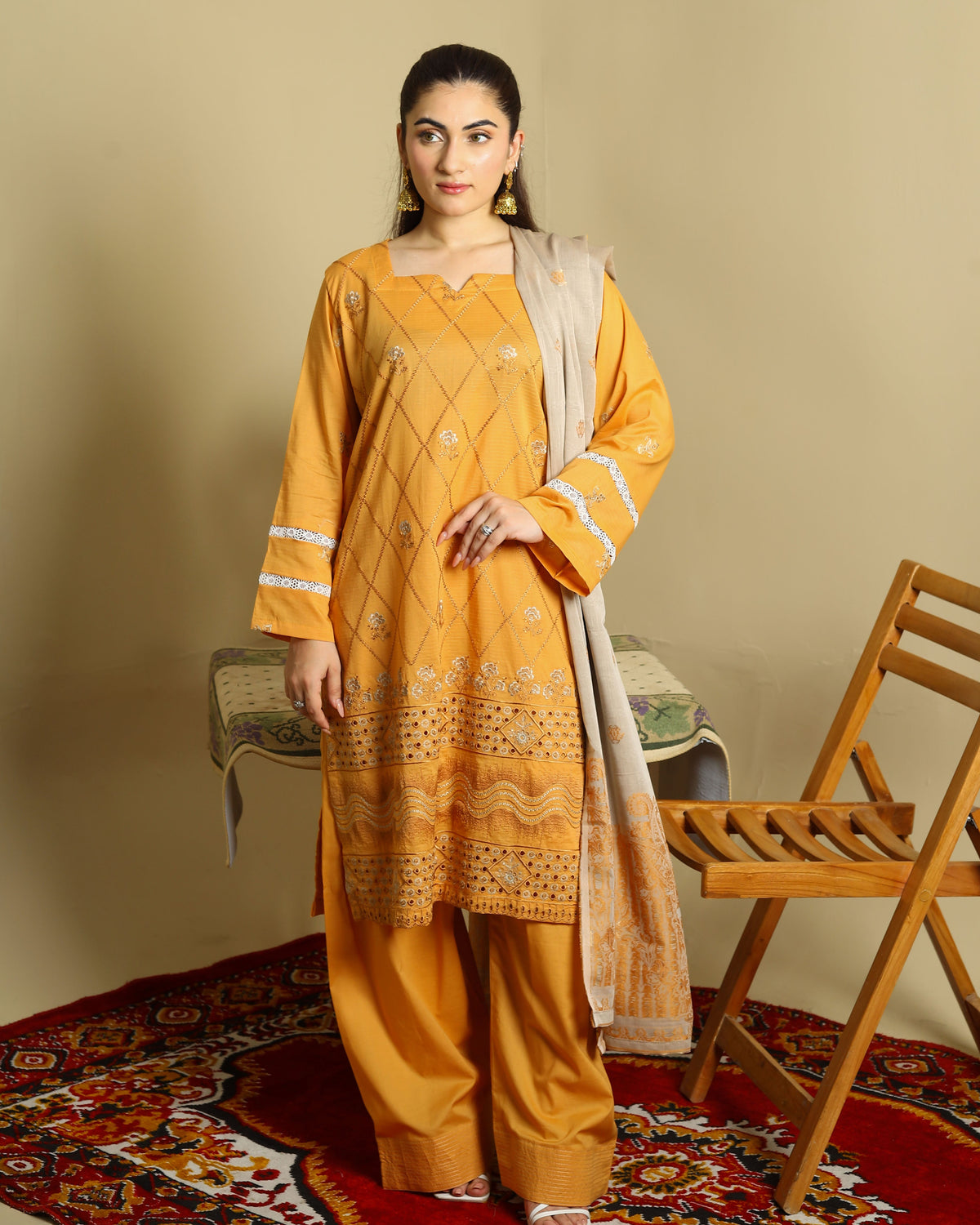 A woman models a mustard yellow ethnic three-piece suit with matching pants and a beige dupatta, standing on a red patterned rug next to a wooden chair against a beige background.