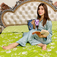 A woman sits on a bed wearing light blue pajamas and holding a mug, with a green and white floral-patterned bedsheet as the main product. The background features an ornate cream-colored headboard and patterned wall decals.