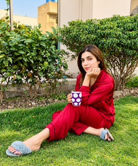 A woman sits on a green lawn wearing a matching red long-sleeve pajama set and blue fuzzy slippers, holding a purple mug with white polka dots. She is positioned outdoors in front of a beige building with green bushes.