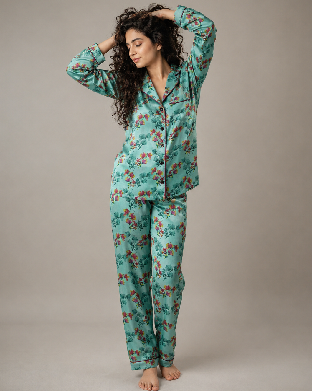 A woman modeling a two-piece pajama set with a long-sleeve button-up top and matching pants, featuring a mint green base with a red and orange floral pattern, set against a plain gray studio background.