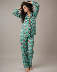 A woman modeling a two-piece pajama set with a long-sleeve button-up top and matching pants, featuring a mint green base with a red and orange floral pattern, set against a plain gray studio background.
